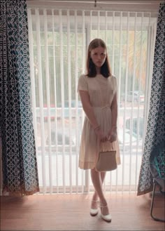 a young woman standing in front of a window holding a purse and looking at the camera