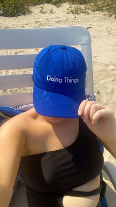 a woman sitting in a chair on the beach wearing a blue hat that says doing things