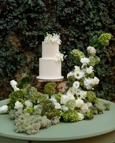 a wedding cake surrounded by flowers and greenery on a table in front of a hedge