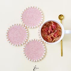 three pink coasters with white pearls and flowers on them next to a spoon, fork and cup