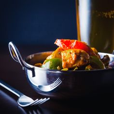 a silver bowl filled with food next to a glass of beer and fork on a table