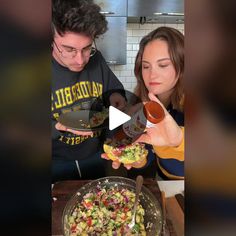 a man and woman standing in front of a bowl of food with a hot dog on it