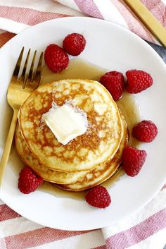 pancakes with butter and raspberries on a white plate next to a knife and fork