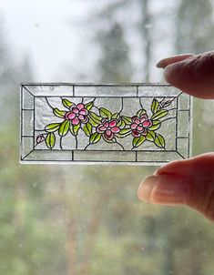 a hand holding up a stained glass window with flowers on it and trees in the background