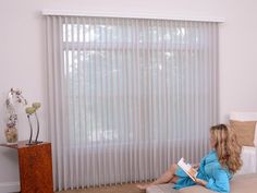 a woman sitting on the floor reading a book in front of a window with vertical blinds