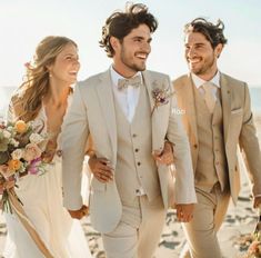 two men and a woman dressed in beige suits walking on the beach with bouquets