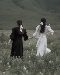 two women in white dresses holding hands and walking through tall grass with mountains in the background