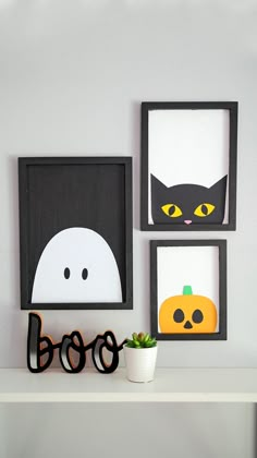 three framed halloween pictures on a shelf with a plant in the foreground and a black cat