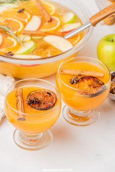 two glasses filled with orange and apple cider next to apples, cinnamon sticks and spices