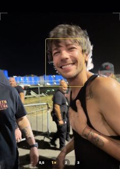 a man with tattoos on his chest standing in front of some people and smiling at the camera