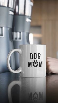 a white coffee mug sitting on top of a table next to a cup with the word dog mom written on it