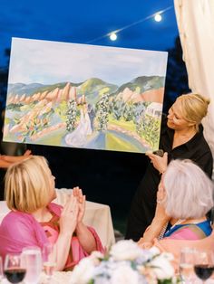 a group of women sitting around a table with paintings on the wall in front of them