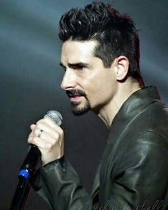 a man with a moustache holding a microphone up to his face while wearing a leather jacket