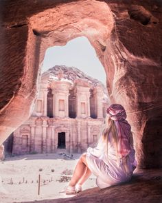 a woman sitting on the side of a cliff looking at an old building in the background