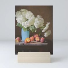 a blue vase filled with white flowers and fruit on top of a wooden block in front of a gray wall