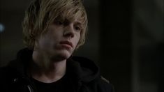 a young man with blonde hair and black jacket looking off into the distance in a dark room