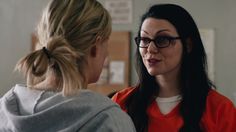 a woman with glasses talking to another woman in an orange shirt and grey sweatshirt,