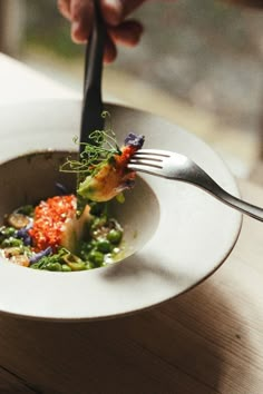 a person is holding a fork in a white bowl with food on the side and garnished with herbs