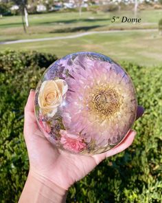 a person holding up a glass ball with flowers on it in front of some bushes