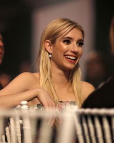 a blonde woman sitting at a table with other people in the background and one smiling