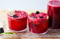 two glasses filled with red liquid and blackberries next to each other on a glass tray