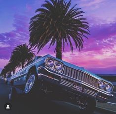 an old car is parked in front of a palm tree at dusk with the sun going down