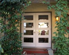 a white double door surrounded by greenery on the side of a house with glass panels