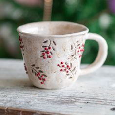 a white cup with red berries on it sitting on top of a wooden table next to a christmas tree