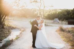 a bride and groom standing on a dirt road in front of some trees at sunset