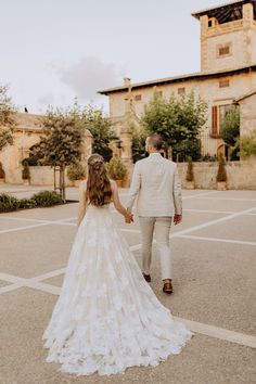 a bride and groom holding hands while walking through an open lot in front of a stone building