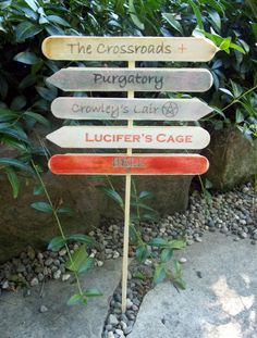 a wooden sign sitting on the side of a road next to some rocks and plants