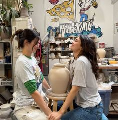 two young women sitting on stools in a pottery shop, one holding a clay pot and the other smiling