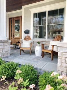 two wooden chairs sitting on top of a stone patio next to flowers and bushes in front of a house