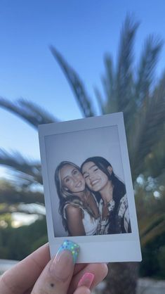 a hand holding up a polaroid with two girls on it and palm trees in the background