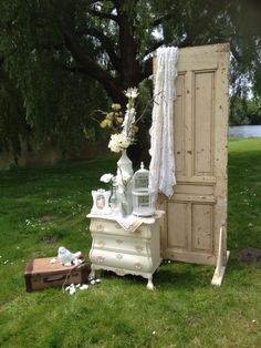 an old dresser with flowers and birdcage next to it on the grass in front of a tree