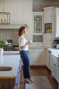 a woman standing in the middle of a kitchen with white cabinets and counter tops, holding a coffee cup