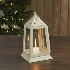 a white lantern with two lit candles in front of a christmas tree on a table