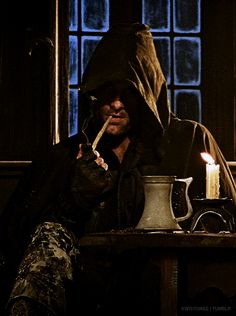 a man sitting at a table with a lit candle in his mouth and wearing a hood