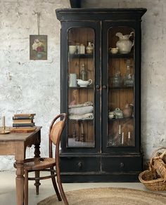 an old fashioned china cabinet with glass doors and shelves in the corner, next to a wicker chair