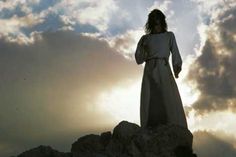 a woman standing on top of a rocky hill under a cloudy sky with the sun behind her