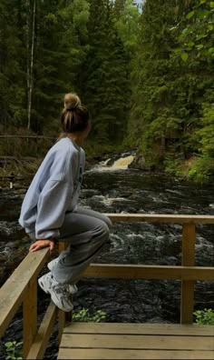 a woman is sitting on a wooden bridge looking at a river in the woods and trees