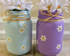 two painted mason jars with daisies tied to them on a tile floor next to a wall