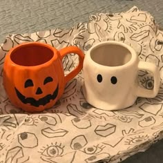 two mugs with faces painted on them are sitting on a bed sheet, one is orange and the other is white