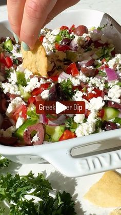 a hand dipping a tortilla chip into a bowl filled with greek salad ingredients