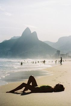 a woman laying on top of a sandy beach next to the ocean in front of mountains