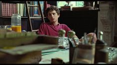 a young man sitting at a table in front of bookshelves and other items