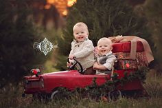 two toddlers sitting on top of a red toy truck in the middle of a forest