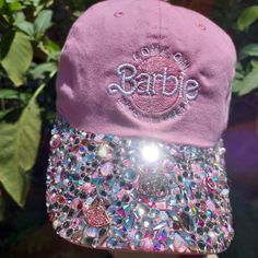 a pink baseball cap with sequins and the word barbie on it, hanging from a tree