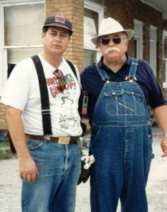 two men standing next to each other in front of a brick building wearing overalls and hats