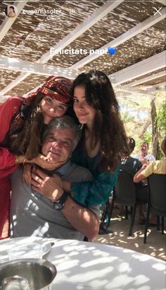 two women and a man are hugging at an outdoor table with people in the background